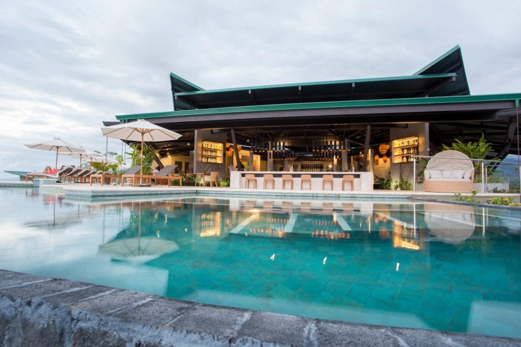A modern open-air bar and lounge area with a row of bar stools faces an inviting swimming pool, surrounded by sun loungers and umbrellas under a cloudy sky.