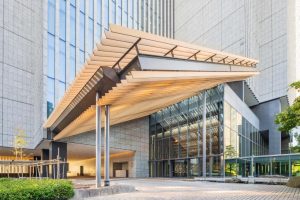 A modern building entrance with a large angular wooden canopy, glass walls, and tall stone facades. The area is paved with stone tiles, and there are some small trees and greenery nearby.