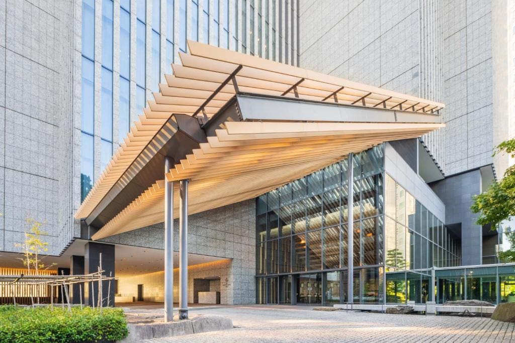 A modern building entrance with a large angular wooden canopy, glass walls, and tall stone facades. The area is paved with stone tiles, and there are some small trees and greenery nearby.
