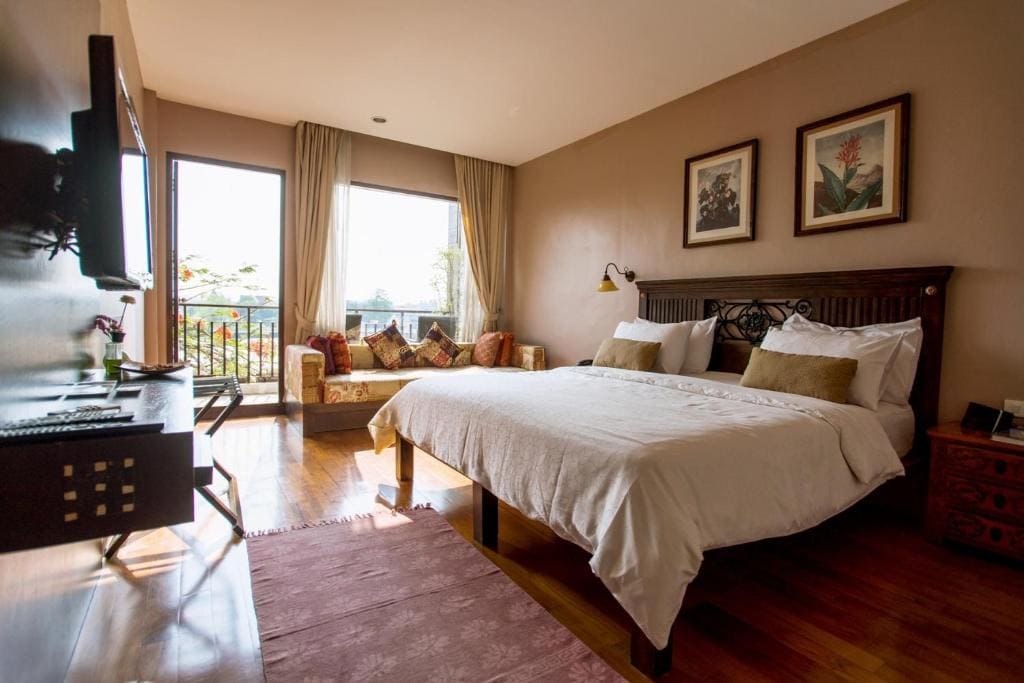 A cozy bedroom with a large bed, decorative pillows, wooden furniture, wall art, a TV, a desk, and large windows letting in natural light, overlooking a balcony with outdoor greenery.