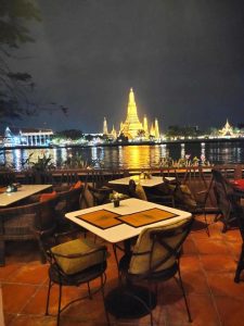 An outdoor restaurant with empty tables and chairs overlooks a river at night, with a brightly illuminated temple, Wat Arun, glowing on the opposite bank under a dark sky.