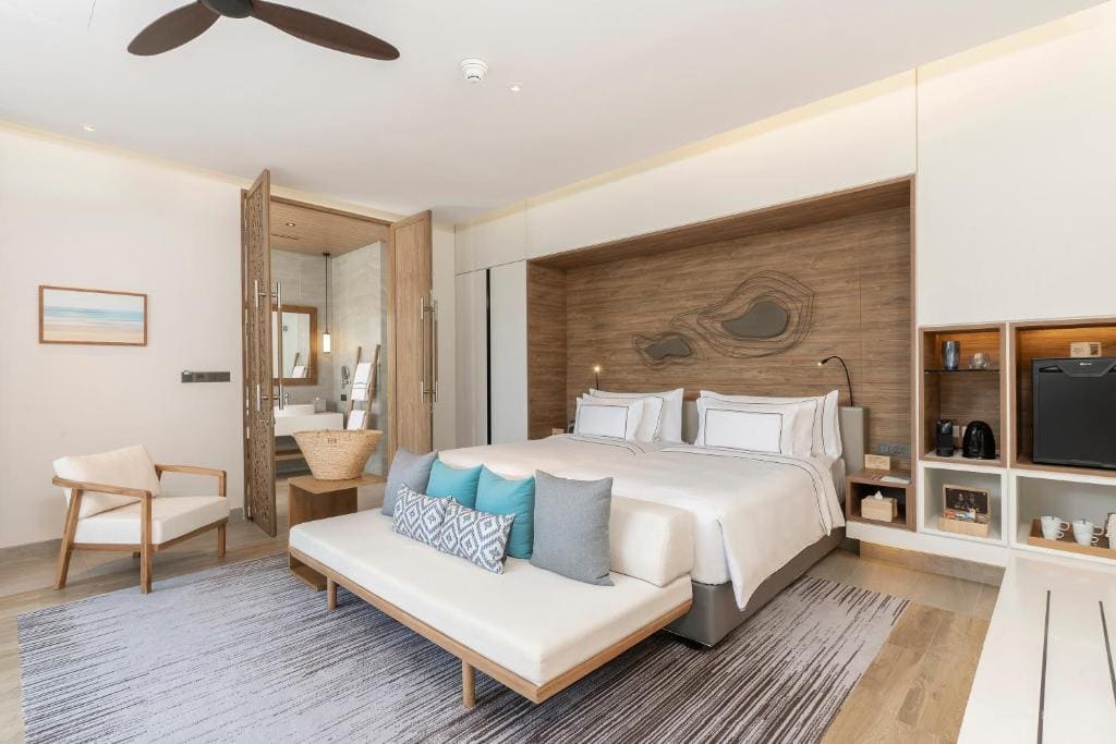 A modern hotel room with a large bed, white linens, and decorative pillows. Theres a cushioned bench at the foot of the bed, a lounge chair, a rug, wall art, shelves with amenities, and an open doorway to the bathroom.