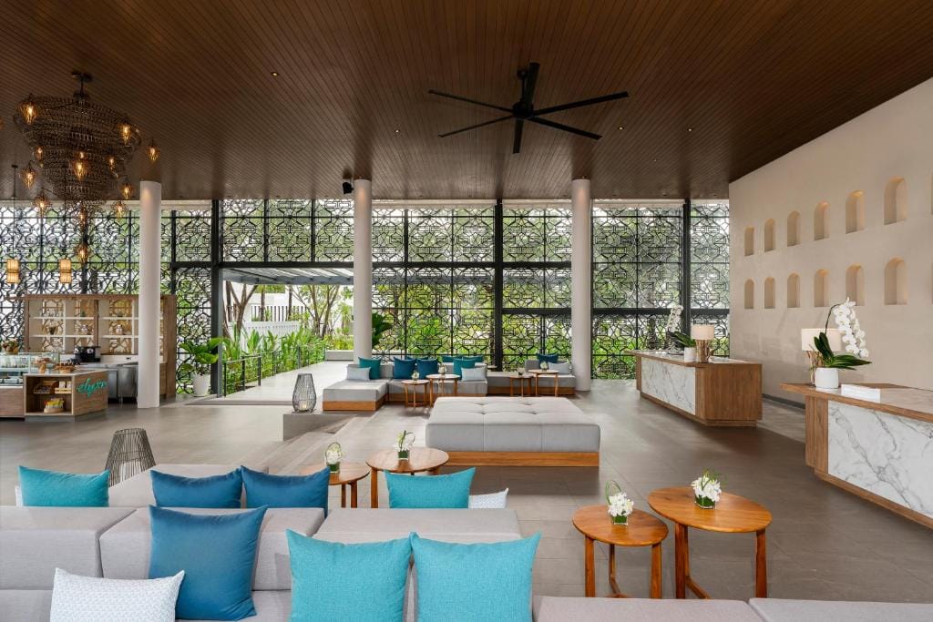 Spacious hotel lobby with modern decor, featuring light sofas with blue and turquoise pillows, wooden tables with small flower arrangements, and large windows with decorative screens overlooking greenery. Reception desks are visible on the right.