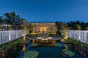 A modern building with a patterned facade is illuminated at dusk, reflecting in a pond with water lilies. The scene is framed by white walls, lush greenery, and palm trees in the background.
