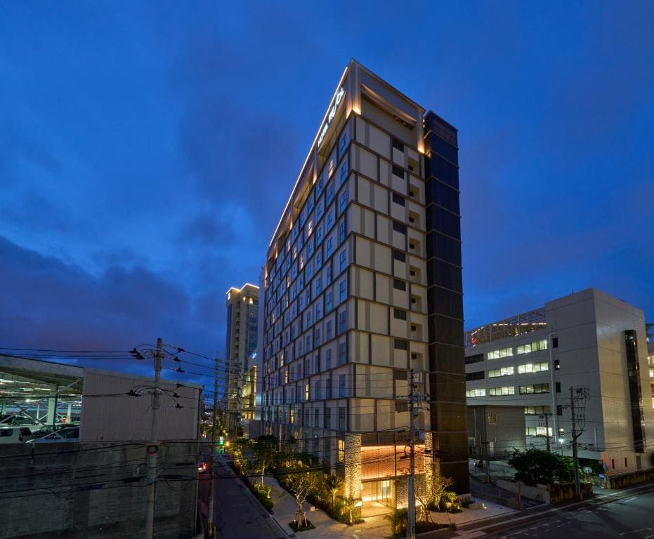 A tall, modern hotel building with many windows is brightly lit at dusk, surrounded by city streets and neighboring structures under a deep blue evening sky.