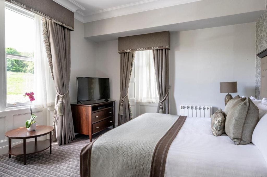 A bright hotel room with a large bed, decorative pillows, a flat-screen TV on a wooden dresser, two windows with long curtains, a small round table with an orchid, and a bedside lamp.