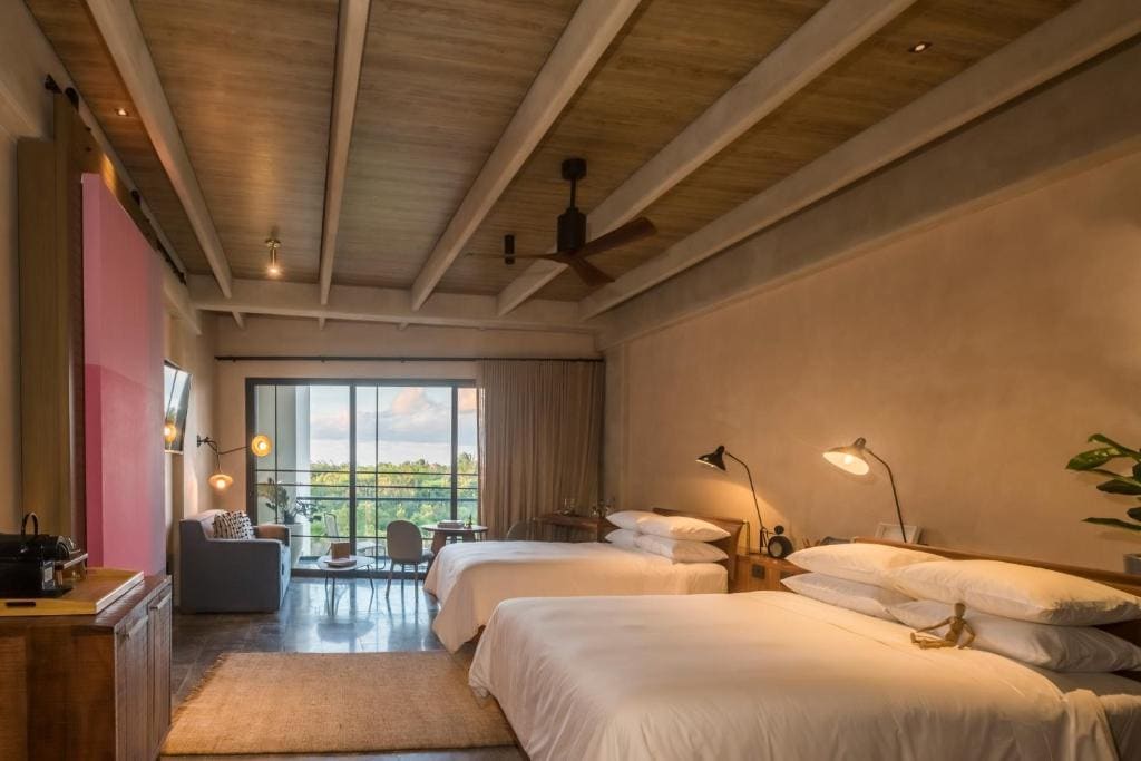 A cozy hotel room with two double beds, modern lighting, wooden ceiling beams, a sitting area near large windows, and a balcony overlooking greenery outside.
