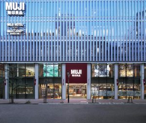 The exterior of a multi-story MUJI store and hotel with large glass windows, displaying products and interiors. The building features MUJI signage in both Japanese and English above the entrance.