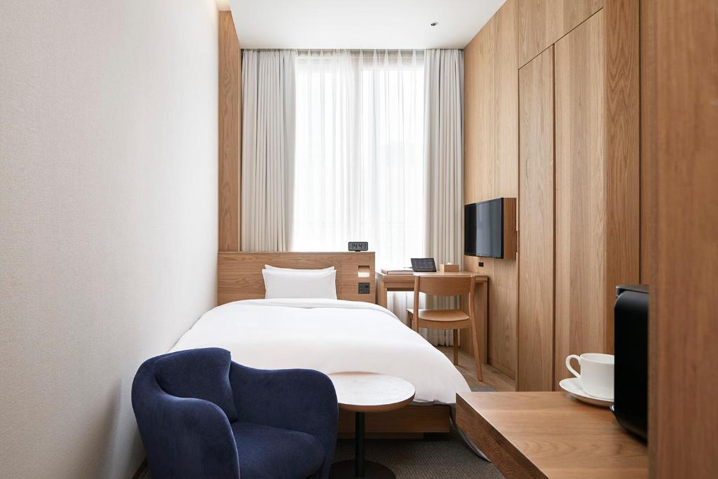 Minimalist hotel room with a single bed, light wood furnishings, a small desk and chair, a wall-mounted TV, a blue armchair, a round side table, and a window with sheer curtains letting in natural light.