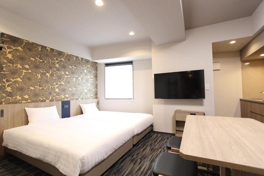 A modern hotel room with two double beds, a floral accent wall, a wall-mounted TV, a light wood table with stools, and a window letting in natural light. The decor features neutral tones and clean lines.