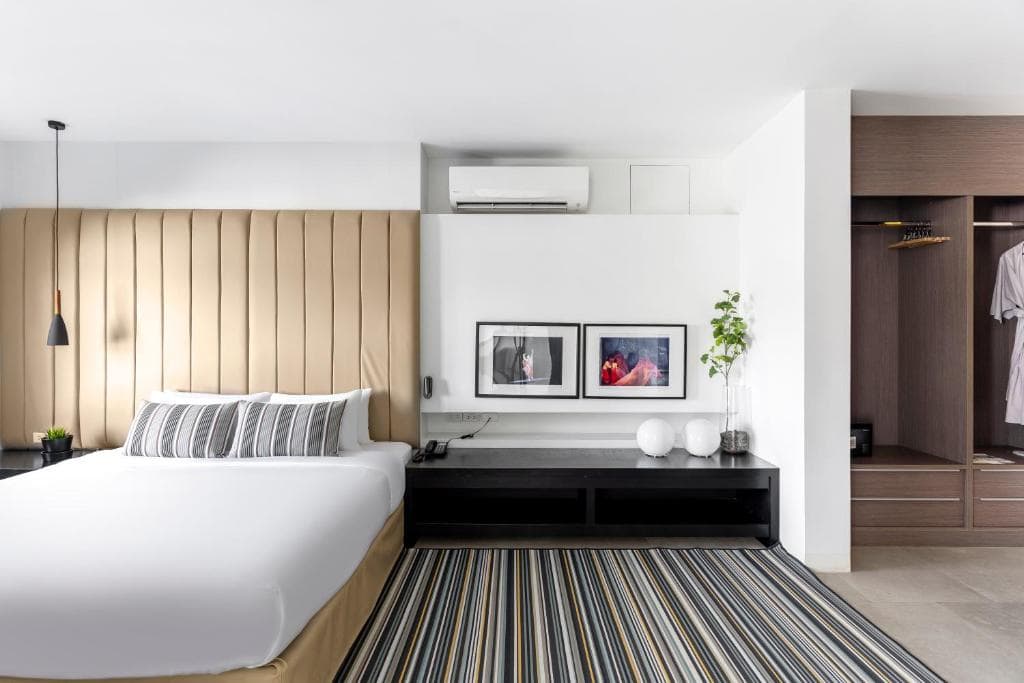 Modern hotel room with a large bed, striped rug, nightstand, open wardrobe with hanging shirts, wall-mounted air conditioner, and framed art above a sleek black console. Decor is minimalist with neutral tones.