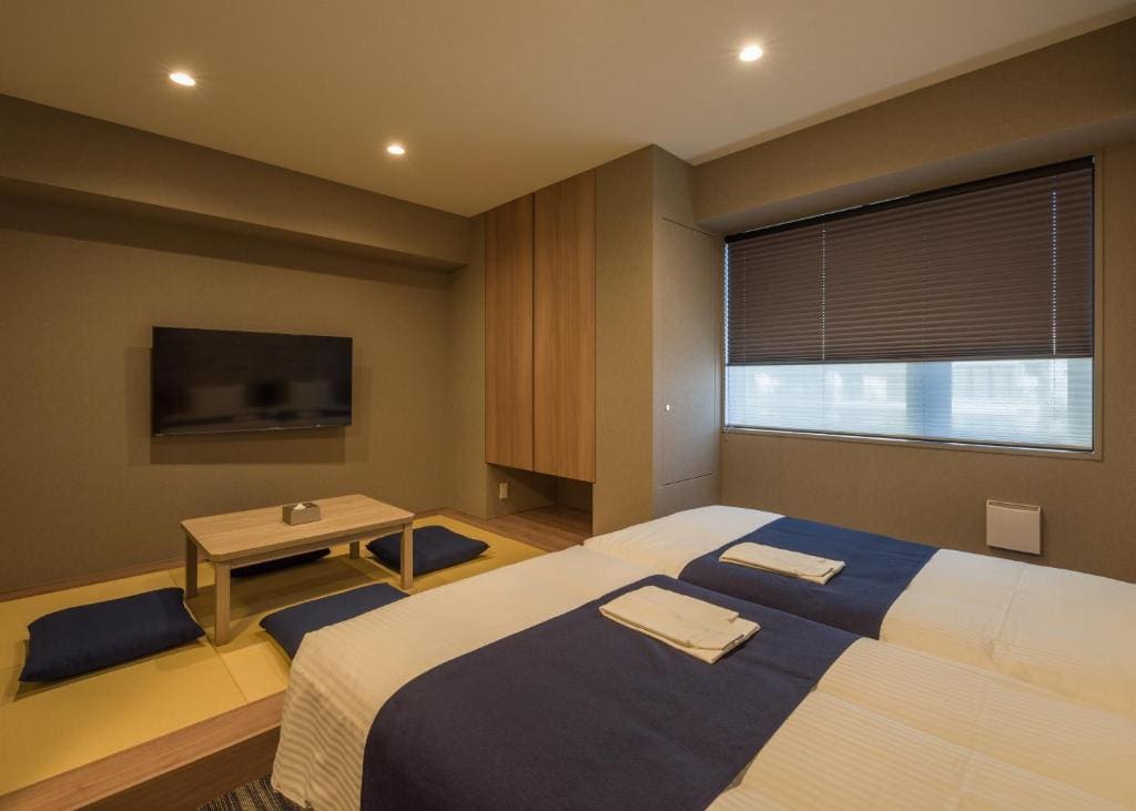 A modern bedroom with two beds covered in white linens and navy throws, a low wooden table with blue floor cushions, a wall-mounted TV, wooden cabinets, and a large window with closed blinds.