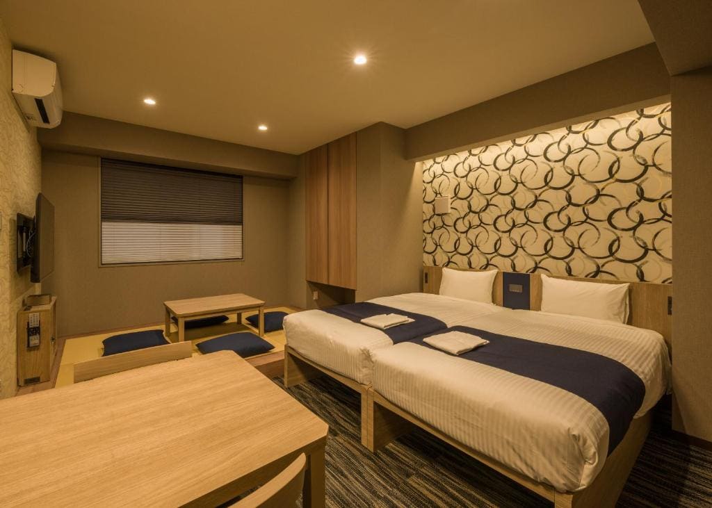 A modern hotel room with two beds, navy blue runners, a wooden table with chairs, a low Japanese-style seating area, wall-mounted TV, and a decorative feature wall with a swirling pattern. Warm lighting creates a cozy atmosphere.