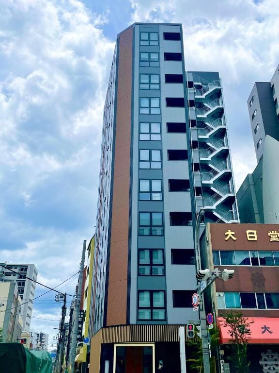 A modern mid-rise building with large windows stands on a city street, surrounded by other buildings under a partly cloudy sky.
