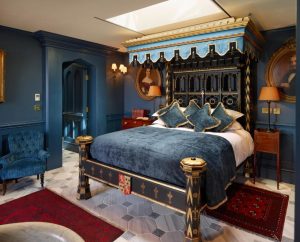 A luxurious bedroom with dark blue walls, a large four-poster canopy bed with gold accents, blue and gold bedding, vintage portraits, red rugs, a blue armchair, and a skylight above the bed.