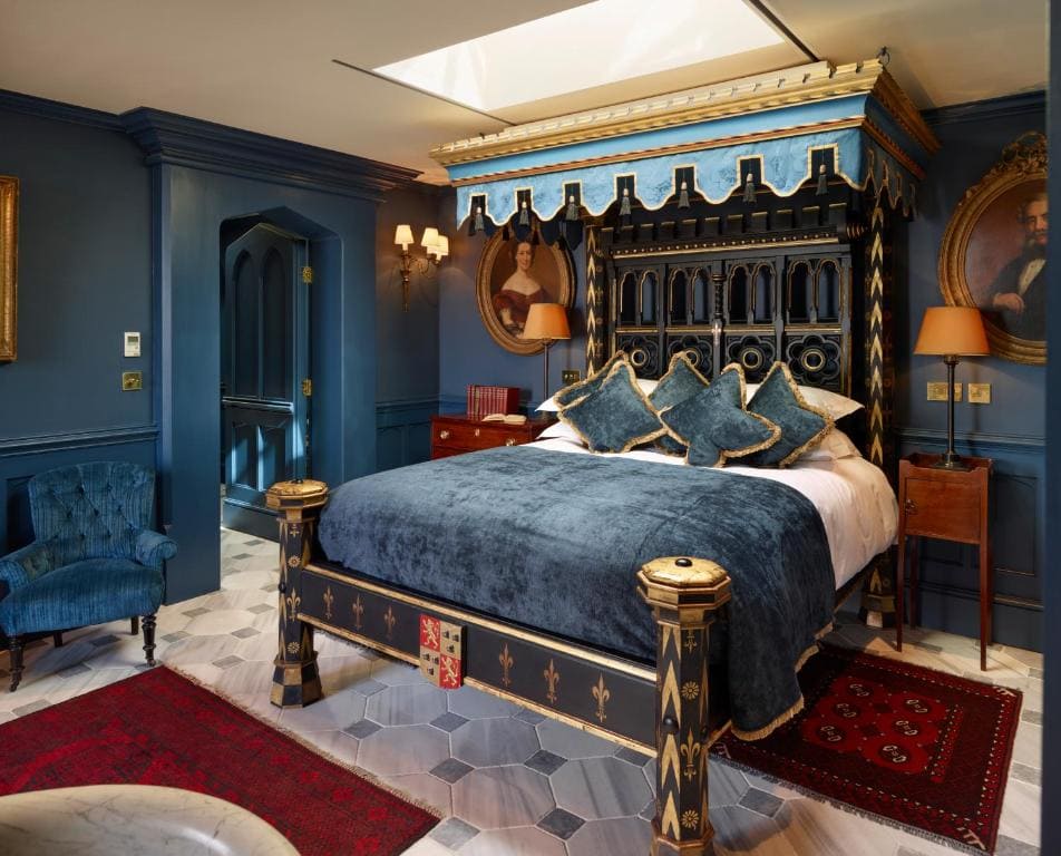 A luxurious bedroom with dark blue walls, a large four-poster canopy bed with gold accents, blue and gold bedding, vintage portraits, red rugs, a blue armchair, and a skylight above the bed.