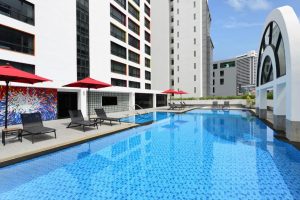 Outdoor swimming pool beside a modern building, with sun loungers and red umbrellas along the poolside. Contemporary mural on the wall, city buildings visible in the background under a clear blue sky.