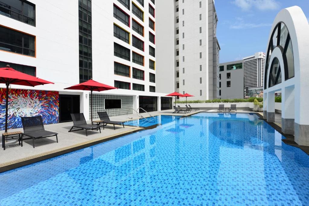 Outdoor swimming pool beside a modern building, with sun loungers and red umbrellas along the poolside. Contemporary mural on the wall, city buildings visible in the background under a clear blue sky.