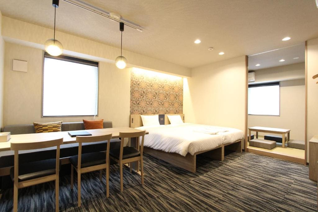 A modern, well-lit hotel room features a double bed with patterned headboard, a dining table with four chairs, a sofa, and a small tatami area with a low table near the window.