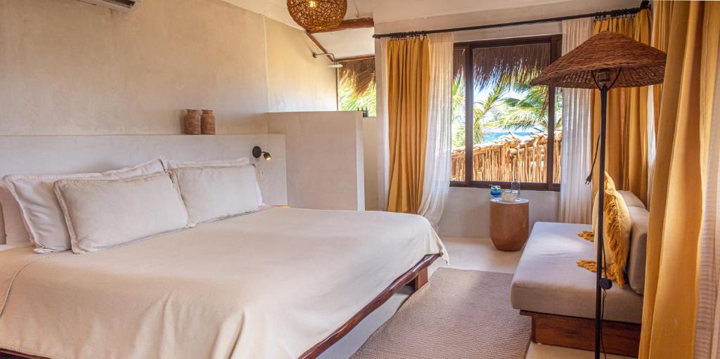 A cozy bedroom with a large bed, white linens, a cushioned bench, warm lighting, and beige curtains. A window shows palm trees and a bamboo fence outside, creating a tropical, relaxing atmosphere.