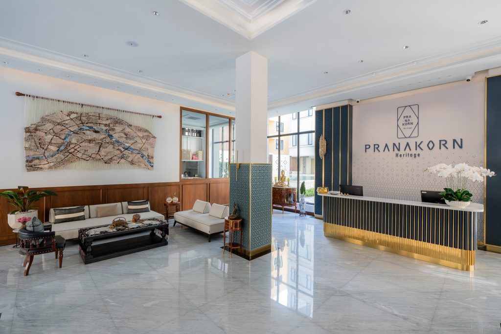 Hotel lobby with modern decor; marble floors, a gold-accented front desk labeled PRANAKORN Heritage, white sofas, a coffee table, a wall tapestry map, and potted plants. Bright natural light fills the room.