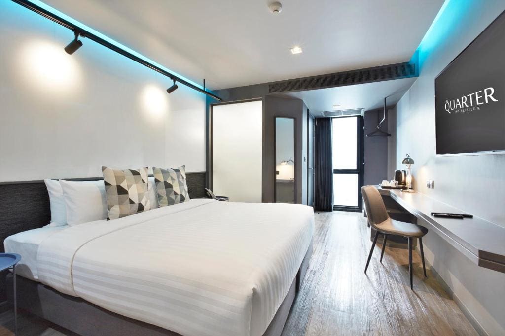 Modern hotel room with a large bed, geometric-patterned pillows, wall-mounted TV, desk with chair, and a door leading to a balcony. The room features minimalist decor and subtle blue accent lighting.