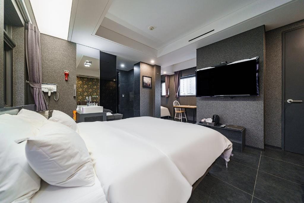 A modern hotel room with a large bed, white linens, gray walls, a flat-screen TV, a small table with chairs, a window with curtains, and a bathroom area with a visible sink and glass shower.