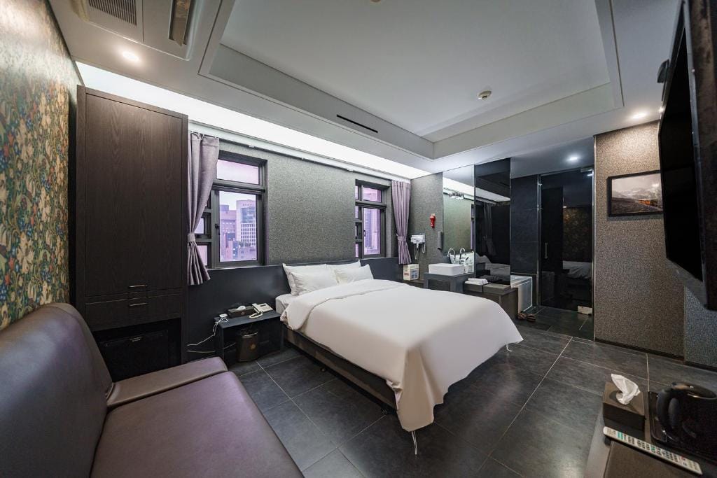 A modern hotel room with a double bed, white bedding, a brown sofa, dark furniture, a wall with floral wallpaper, large windows with city views, and an open bathroom area with a sink and mirror.