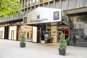 Entrance of the Millennium Hotel in London, with a silver canopy, two potted plants on either side, and large glass windows displaying mannequins in a shop next door.