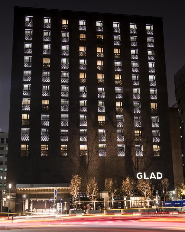 A tall, modern hotel building at night with illuminated windows and the word GLAD displayed on its facade. Light trails from passing cars are visible in front of the building.