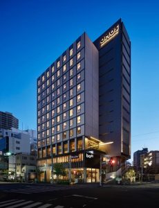 A modern, tall hotel building is brightly lit against a deep blue evening sky, with illuminated windows and signs for OMO3 and shops at street level. The surrounding area has smaller buildings and quiet streets.
