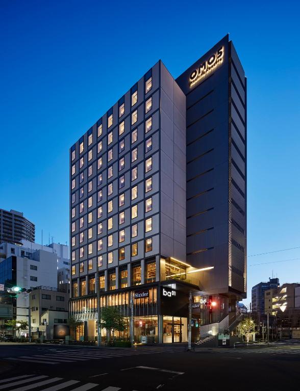 A modern, tall hotel building is brightly lit against a deep blue evening sky, with illuminated windows and signs for OMO3 and shops at street level. The surrounding area has smaller buildings and quiet streets.