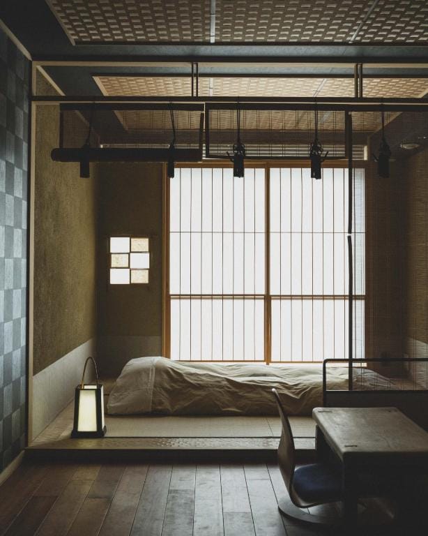 A minimalist Japanese-style room with a low futon bed on a tatami mat, a wooden desk and chair, shoji screen windows letting in light, and a modern lantern on the floor. The ambiance is calm and serene.