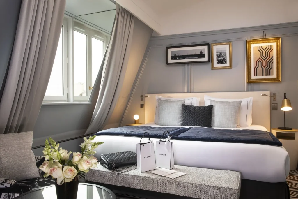 A stylish hotel room features a large bed with gray and white bedding, elegant pillows, framed art on the wall, a window with gray curtains, fresh flowers, and Chanel shopping bags on a table.
