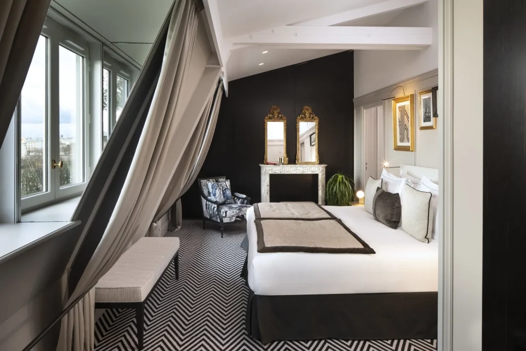 Modern hotel room with a king-sized bed, chevron-patterned carpet, elegant mirrors above a marble fireplace, armchair, large windows with curtains, and framed art on dark accent walls.