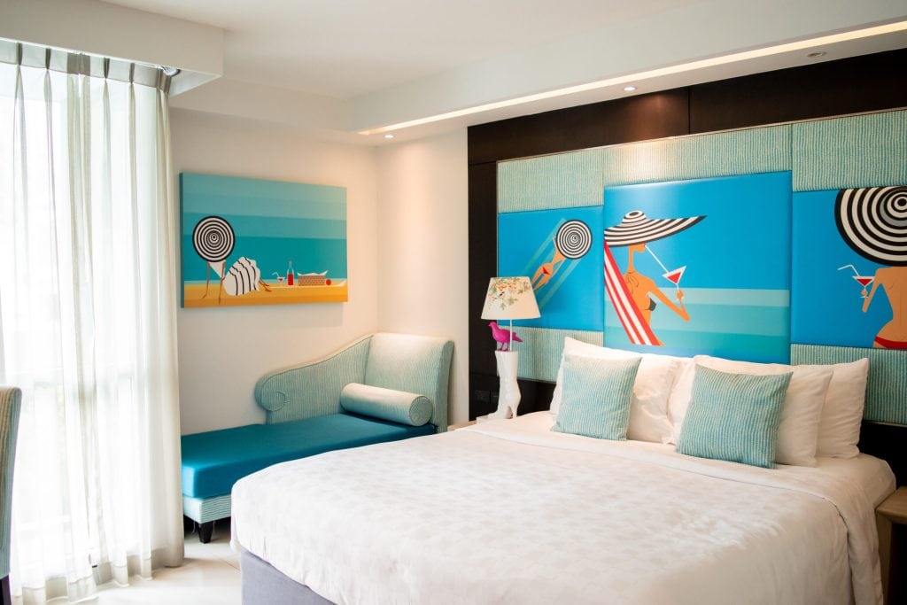 Bright hotel room with a large bed, white linens, aqua blue pillows, a blue chaise lounge, and wall art depicting beach scenes with women in sun hats and umbrellas. Sunlight streams in through sheer curtains.