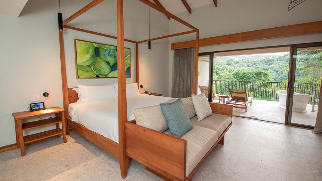 Spacious bedroom with a wooden canopy bed, white bedding, a sofa at the foot of the bed, nightstands, and large sliding doors opening to a balcony with a view of trees and hills.
