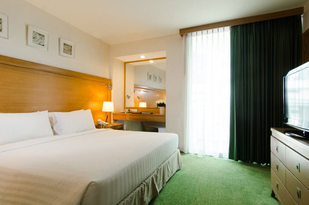 A bright hotel room with a large bed, white linens, a wooden headboard, bedside lamp, desk with mirror, green carpet, and a window with sheer curtains allowing natural light in.