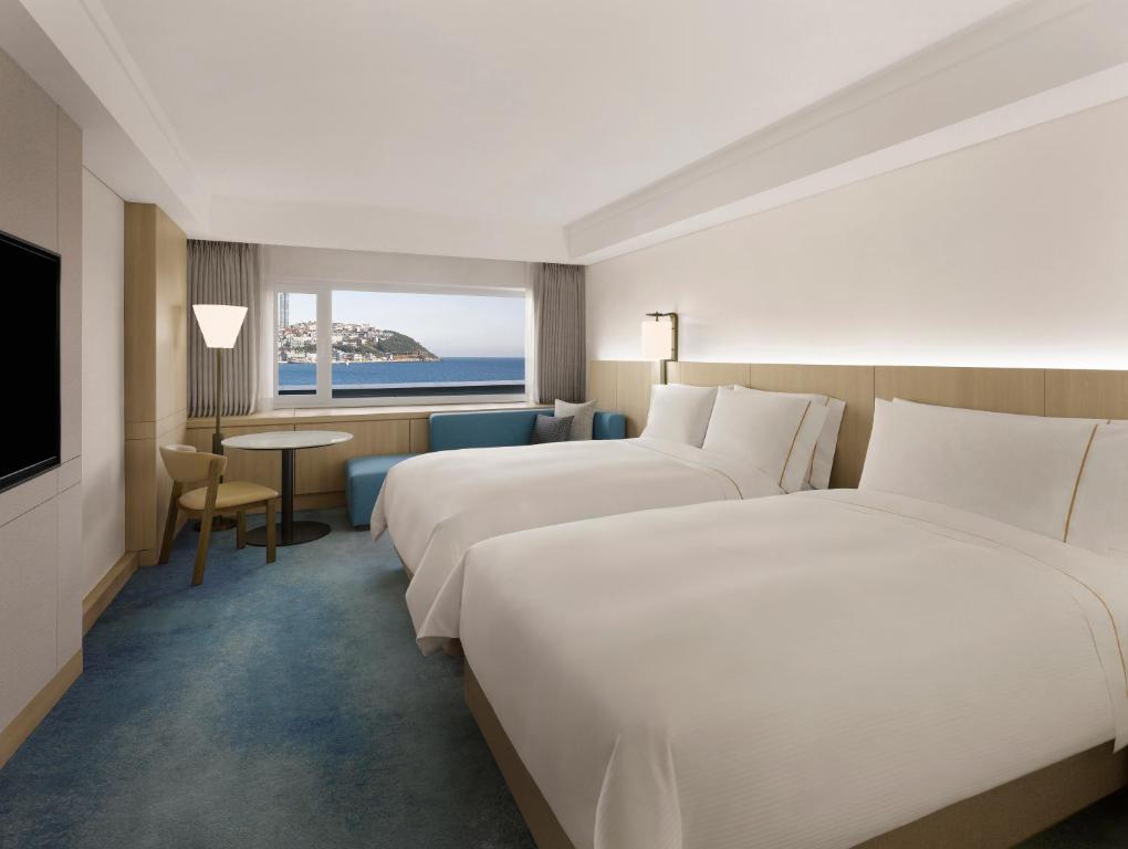 Modern hotel room with two large beds, a flat-screen TV, a small round table with two chairs, a window seat, and a view of the ocean and coastline through a wide window. The decor is light and minimalist.