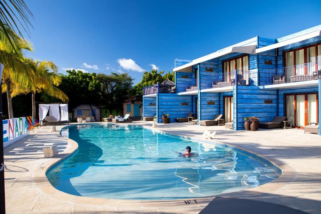 A person swims in a clear, curved pool surrounded by lounge chairs at a modern, blue-painted hotel with balconies and palm trees under a bright blue sky.