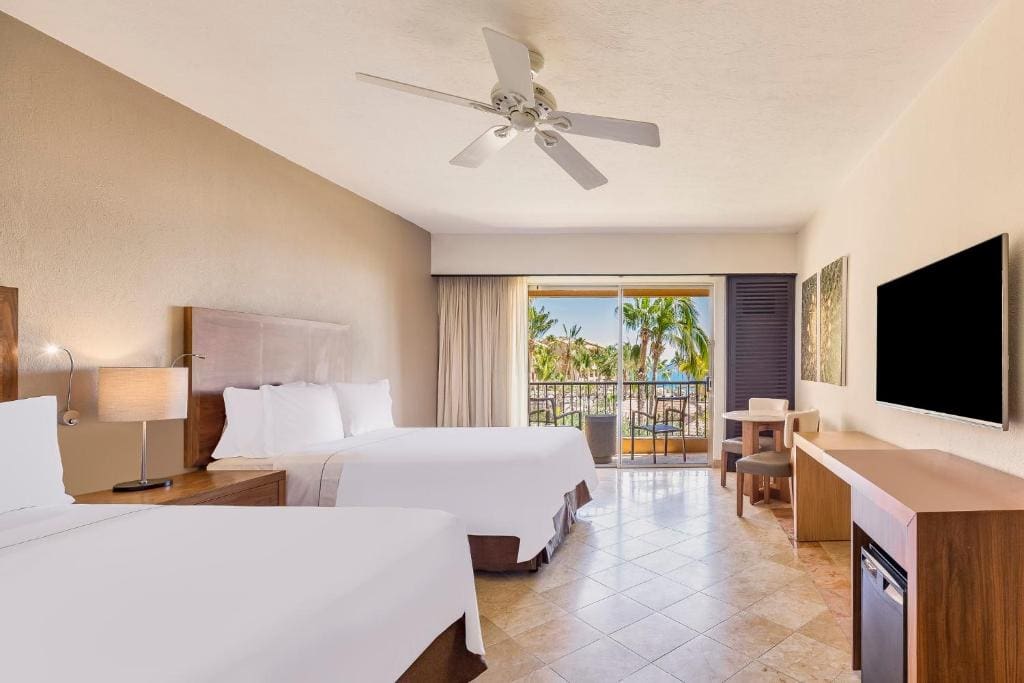 A bright hotel room with two double beds, a ceiling fan, a wall-mounted TV, a desk, and a balcony overlooking palm trees and greenery outside. Natural light fills the space through sliding glass doors.