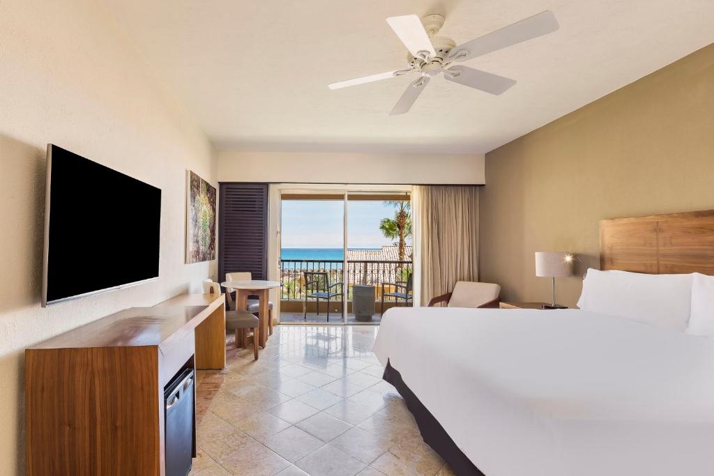 A modern hotel room with a large bed, TV, desk, mini fridge, and chair. Sliding glass doors open to a balcony with a view of the ocean, palm trees, and blue sky.