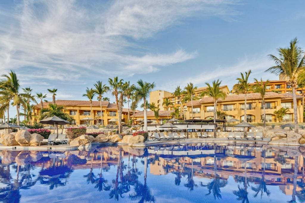 Luxury resort with palm trees and multiple buildings reflected in a large, blue swimming pool under a partly cloudy sky, with lounge chairs and umbrellas around the pool area.