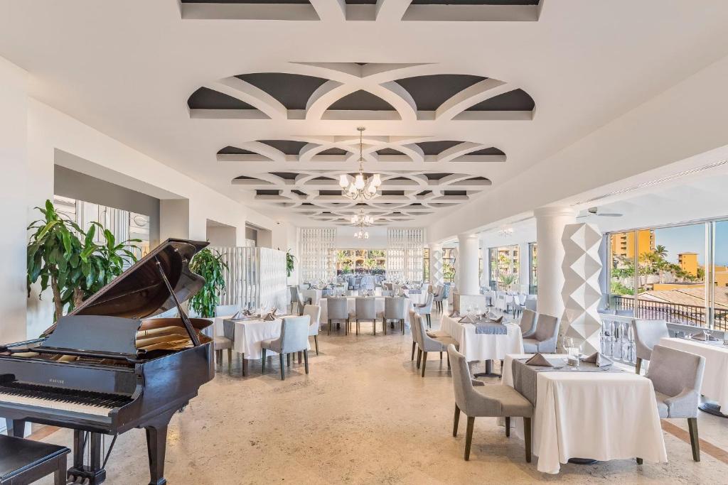 Elegant restaurant interior with white walls and arched ceiling details, set with white tablecloths and gray chairs. A grand piano sits in the foreground, and large windows let in abundant natural light.