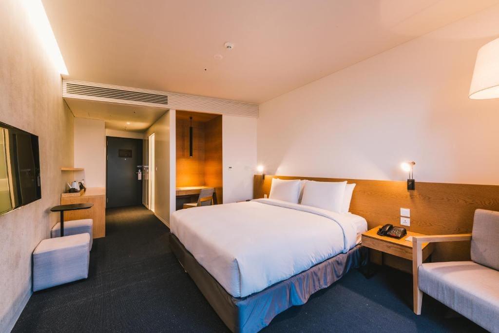 A modern hotel room with a large bed, bedside tables, reading lamps, a wall-mounted TV, a desk with a chair, and a comfortable armchair. The room has soft lighting and a minimalist design.