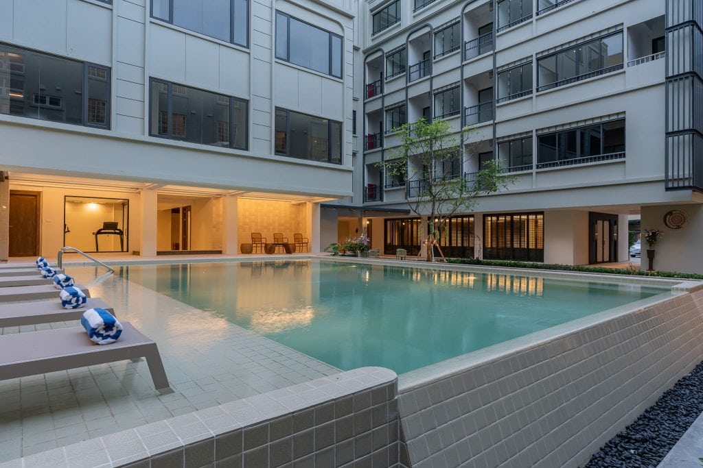A modern outdoor pool surrounded by beige tiles, with lounge chairs and rolled towels along one side, set in the courtyard of a multi-story building with large windows.