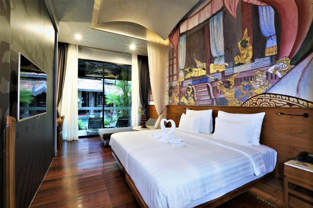 A hotel room with a large bed, white linens, and towel swans. A colorful mural decorates the wall above the bed. Floor-to-ceiling windows reveal greenery outside, creating a bright and inviting atmosphere.