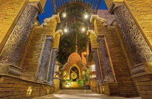Ornate stone archways with intricate carvings frame a pathway leading to a golden gong in a lush, warmly-lit courtyard, surrounded by trees and decorated with lanterns at twilight.