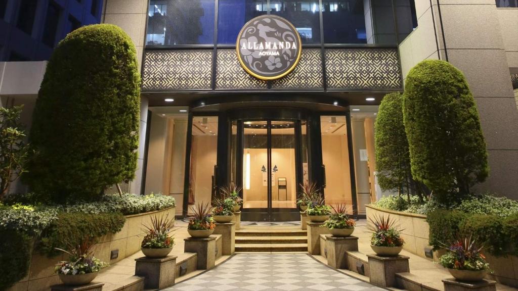 Entrance of the Allamanda Hotel at night, with round double glass doors, manicured shrubs, potted plants, steps leading up, and a large round sign above the entrance displaying the hotel name.