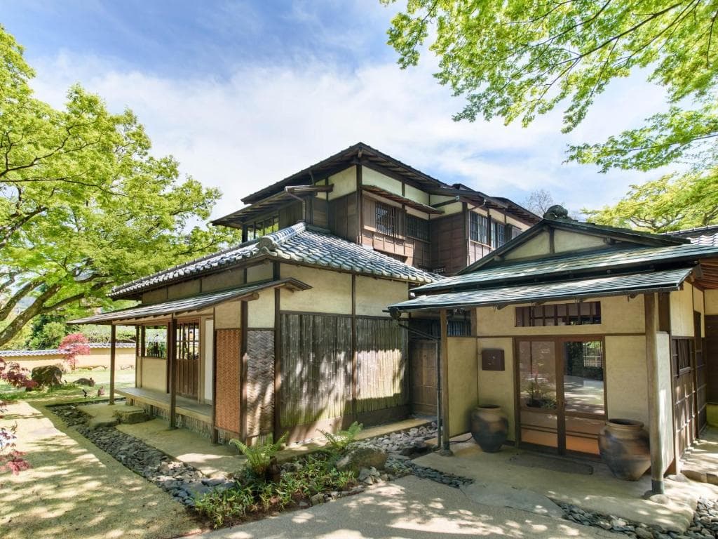 Traditional Japanese house with wooden and plaster walls, tiled roof, and sliding doors, surrounded by lush green trees and a landscaped garden under a bright blue sky.